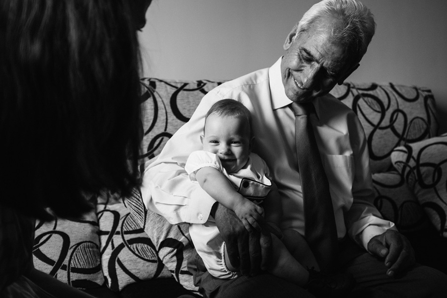 fotos de bautizo, el abuelo sentado en el sillon con su nieto en brazos, el niño sonrie a su tia